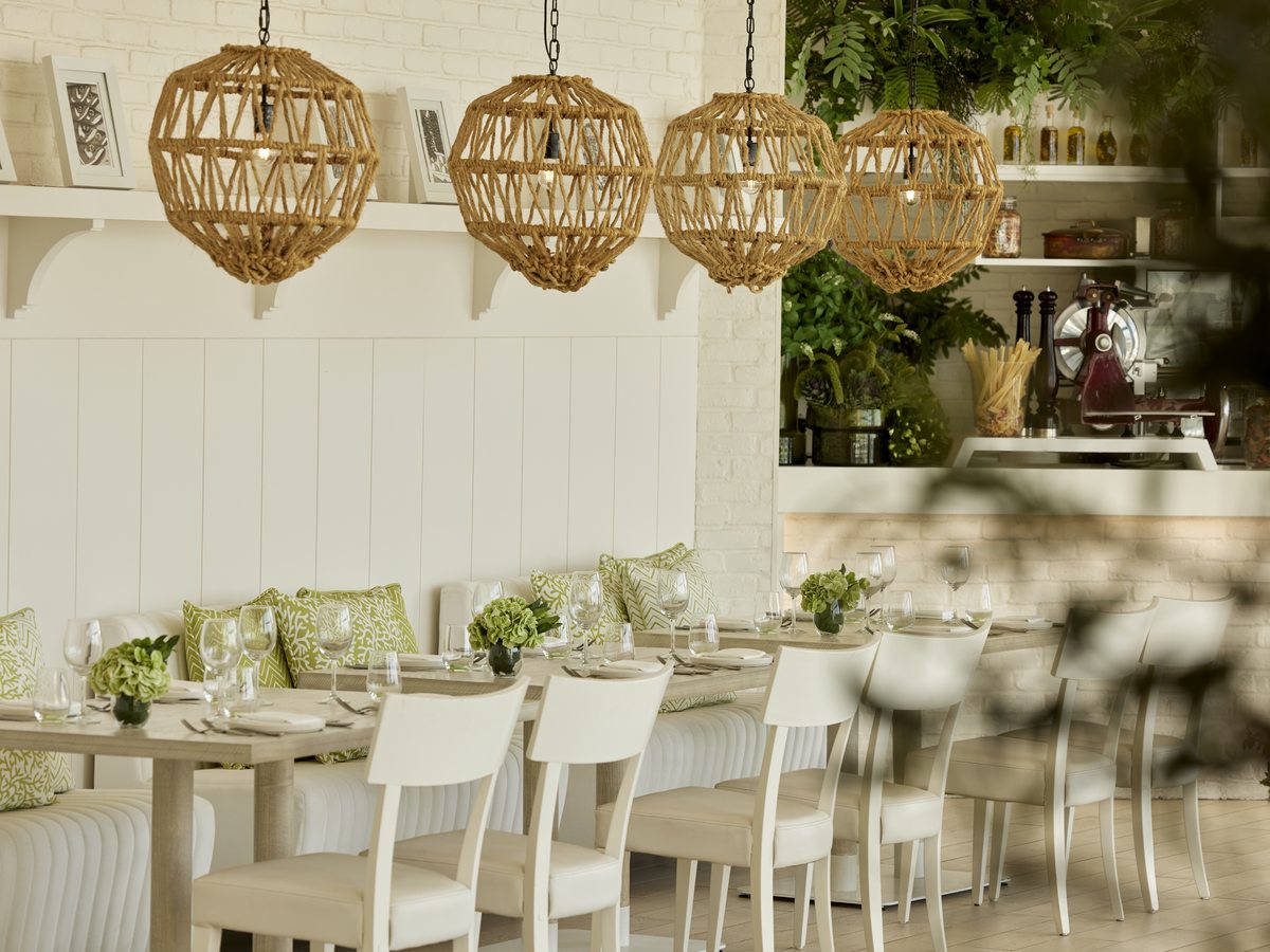 The white panelled dining room at Fresco, with woven orb pendant lights, a cushioned banquette and a planted open kitchen.