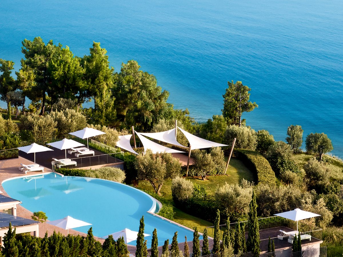 A curved pool on a garden terrace with white shade sails and loungers, pines and cypresses around it and deep blue sea below.