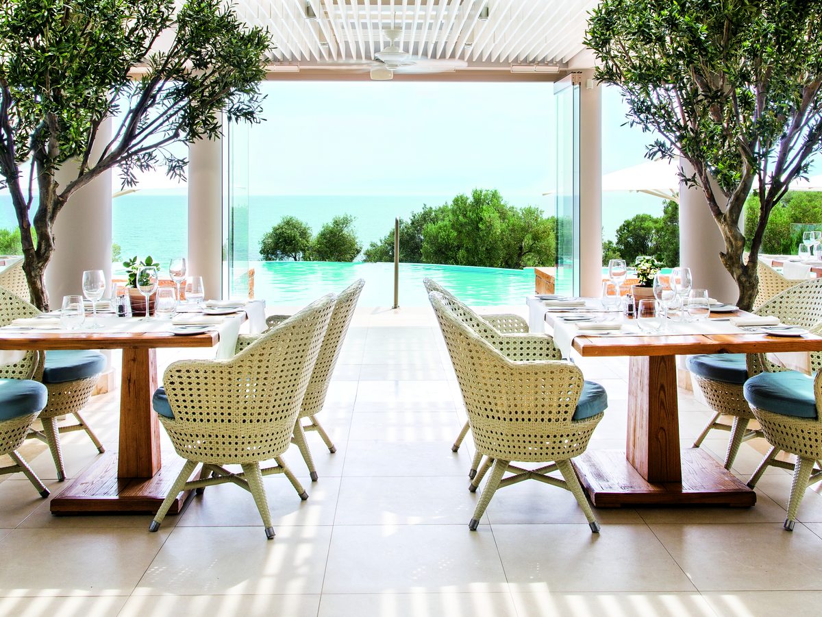 Tables with woven armchairs under a white pergola at Provence, opening onto olive trees, the pool and the sea.