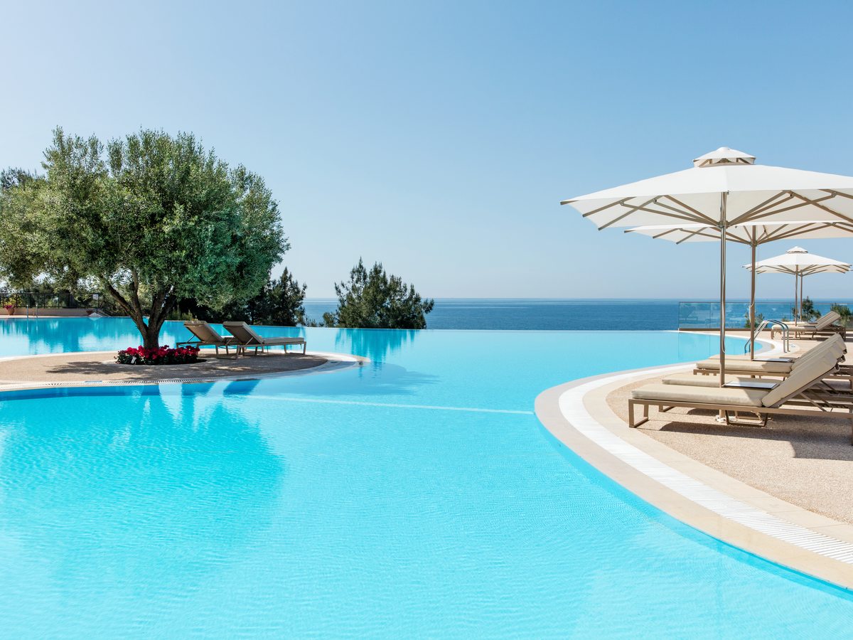 The main infinity pool with an olive tree on a planted island, sun loungers and a white parasol at the water’s edge and the sea behind.