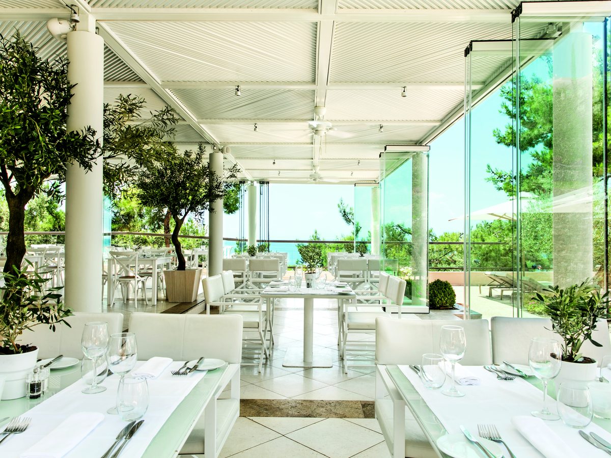 The glass-walled dining pavilion at Ouzo, with white linen tables, olive trees planted in the room and the sea through the far wall.