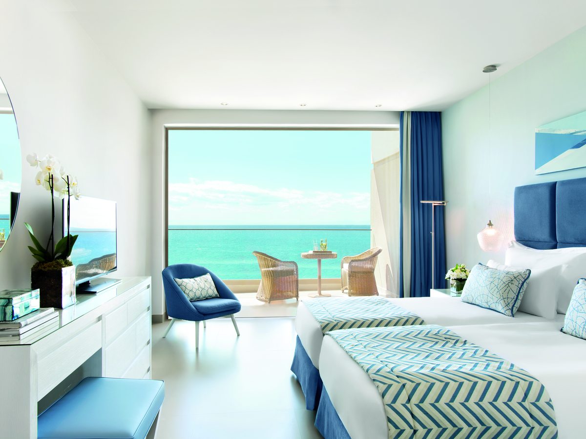 A Family Room with twin beds, a blue armchair and a dressing table, and a wide window onto a terrace and the sea.