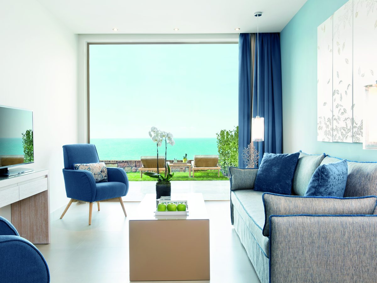 The living room of a One Bedroom Family Suite, with a sofa, blue armchairs, a coffee table and glass doors onto the garden and the sea.