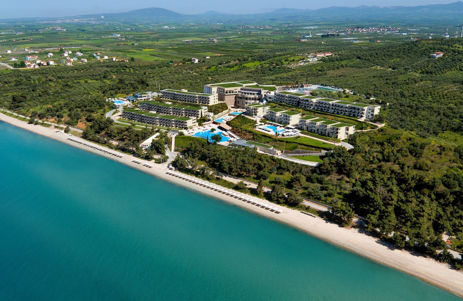 Ikos Oceania from the air: terraced room blocks rising up a green hillside above a sandy beach and the Aegean.