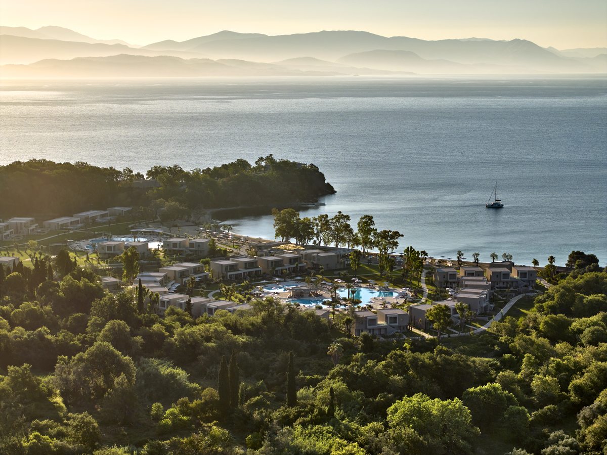 Aerial view of the low white resort buildings among pine, citrus and olive on a green headland, with the calm blue bay beyond.