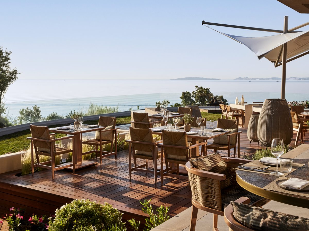 The open deck at Elia, set with wooden tables, woven chairs and a parasol, the sea and hills in the distance.