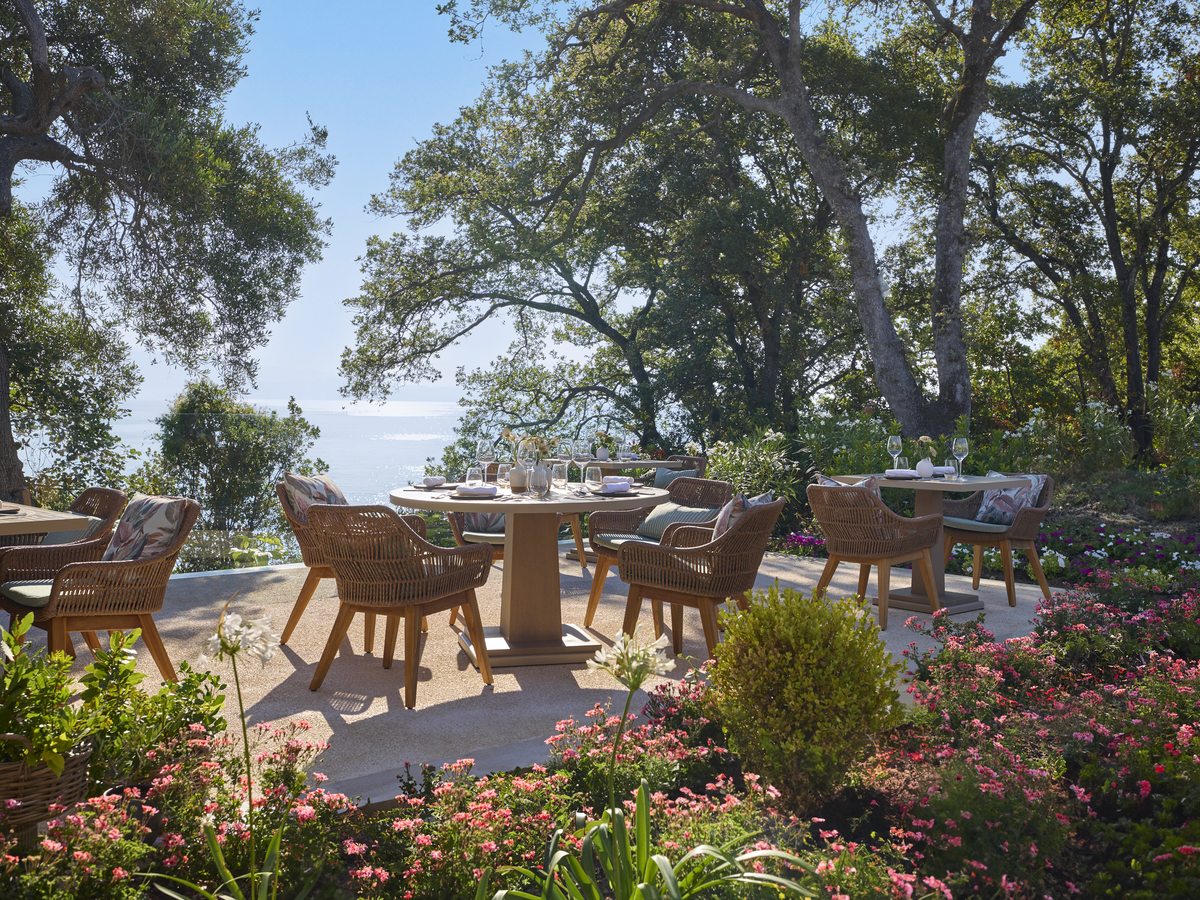 Round tables and rattan chairs on a paved terrace among trees and flowering shrubs, with the sea beyond.