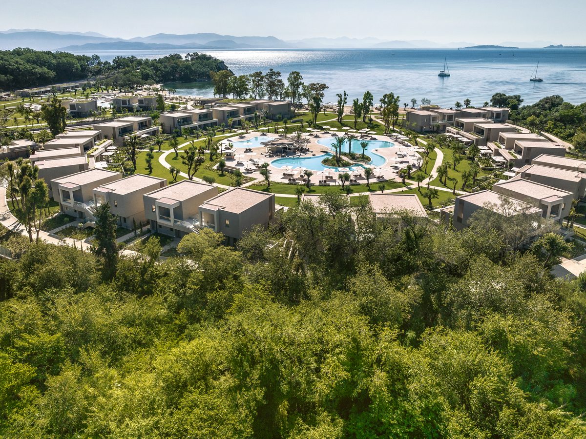 Aerial view of low-rise bungalow blocks set around a large curved pool, with lawns, paths and trees between them.