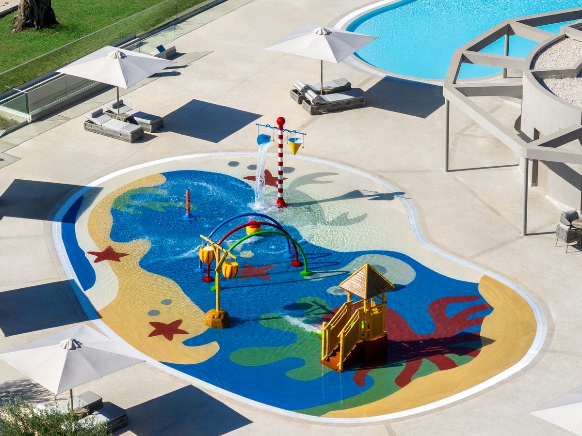 A shallow splash park painted with sea creatures, with water jets, small slides and shaded loungers around it.