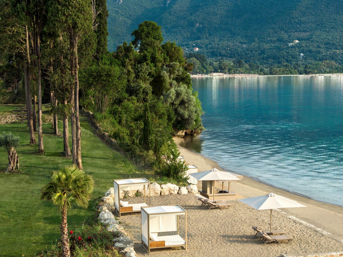 Canopied daybeds and parasols set out on the sand at the resort beach, with the wooded far side of the bay across the water.