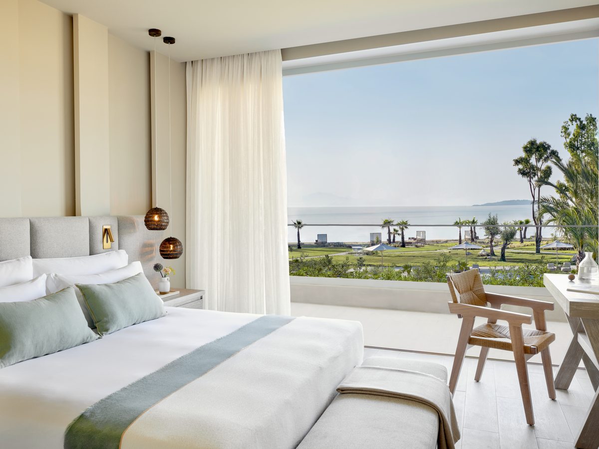 A bungalow suite bedroom with a wide window and a furnished balcony, palms and the sea in the distance.