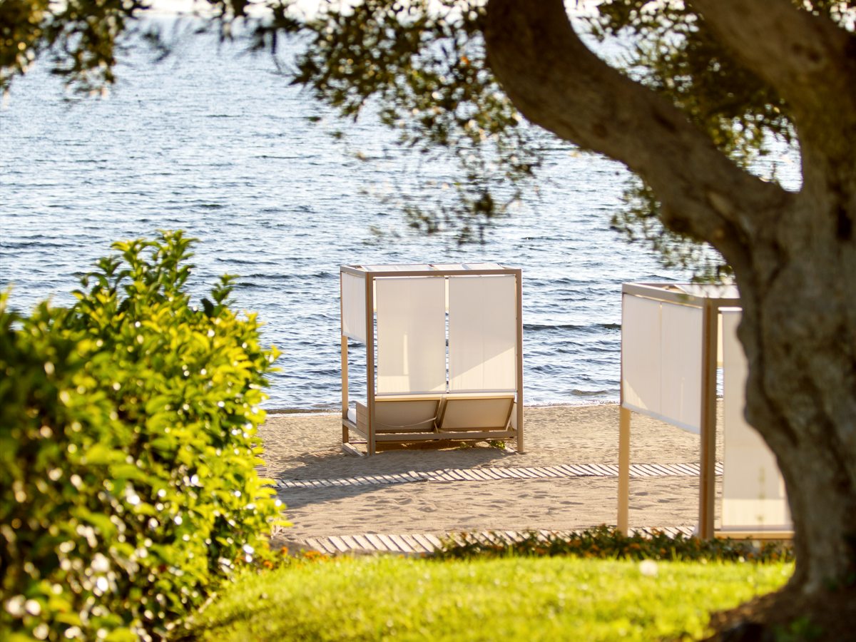 Two curtained canopy daybeds standing on the sand at the edge of the sea, seen past an olive tree.