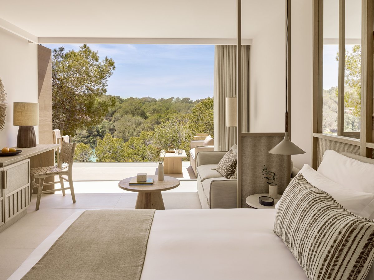A Junior Suite with a bed and a sofa opening through full-height glass onto a terrace, with a pool and trees beyond.