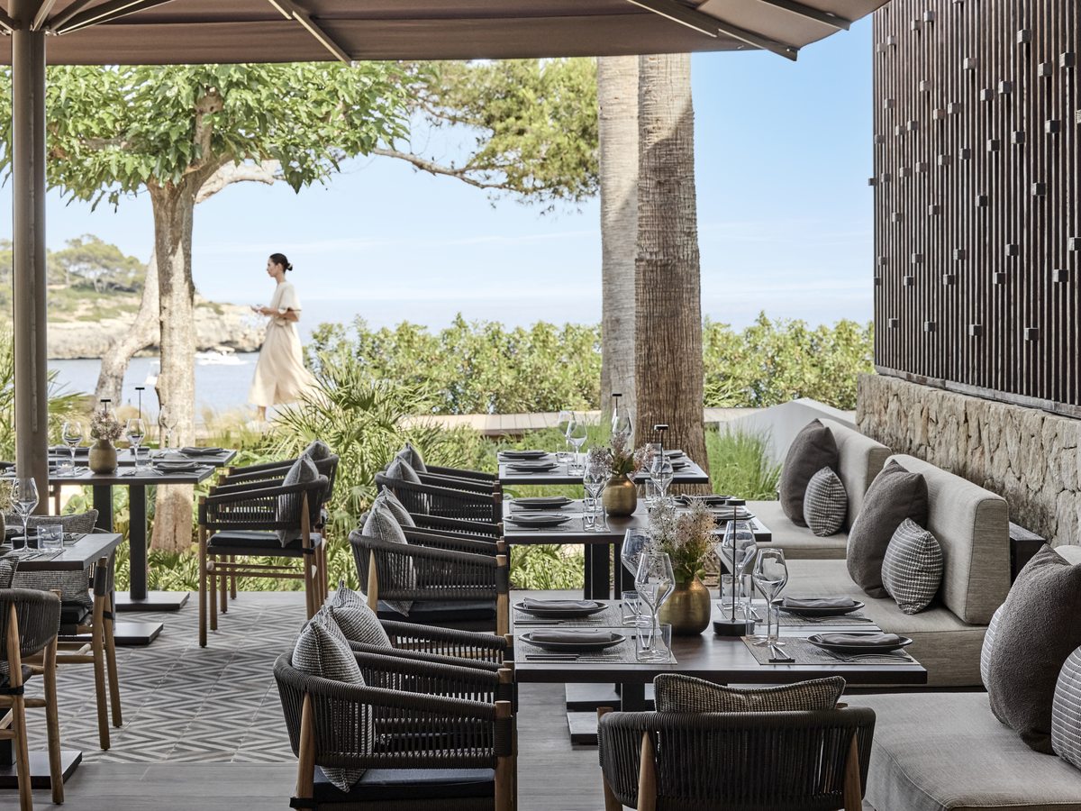 The terrace at Anaya, dark rattan chairs and cushioned banquettes around set tables under a canopy, with pines and the sea beyond.
