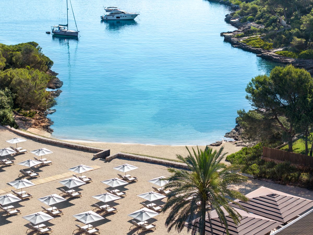 Rows of sun loungers and parasols on the sand at the resort cove, with boats moored on the turquoise water.