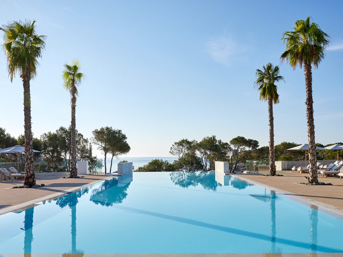 A long pool with a flush edge, ringed by tall palms and rows of sun loungers, with the sea on the horizon.