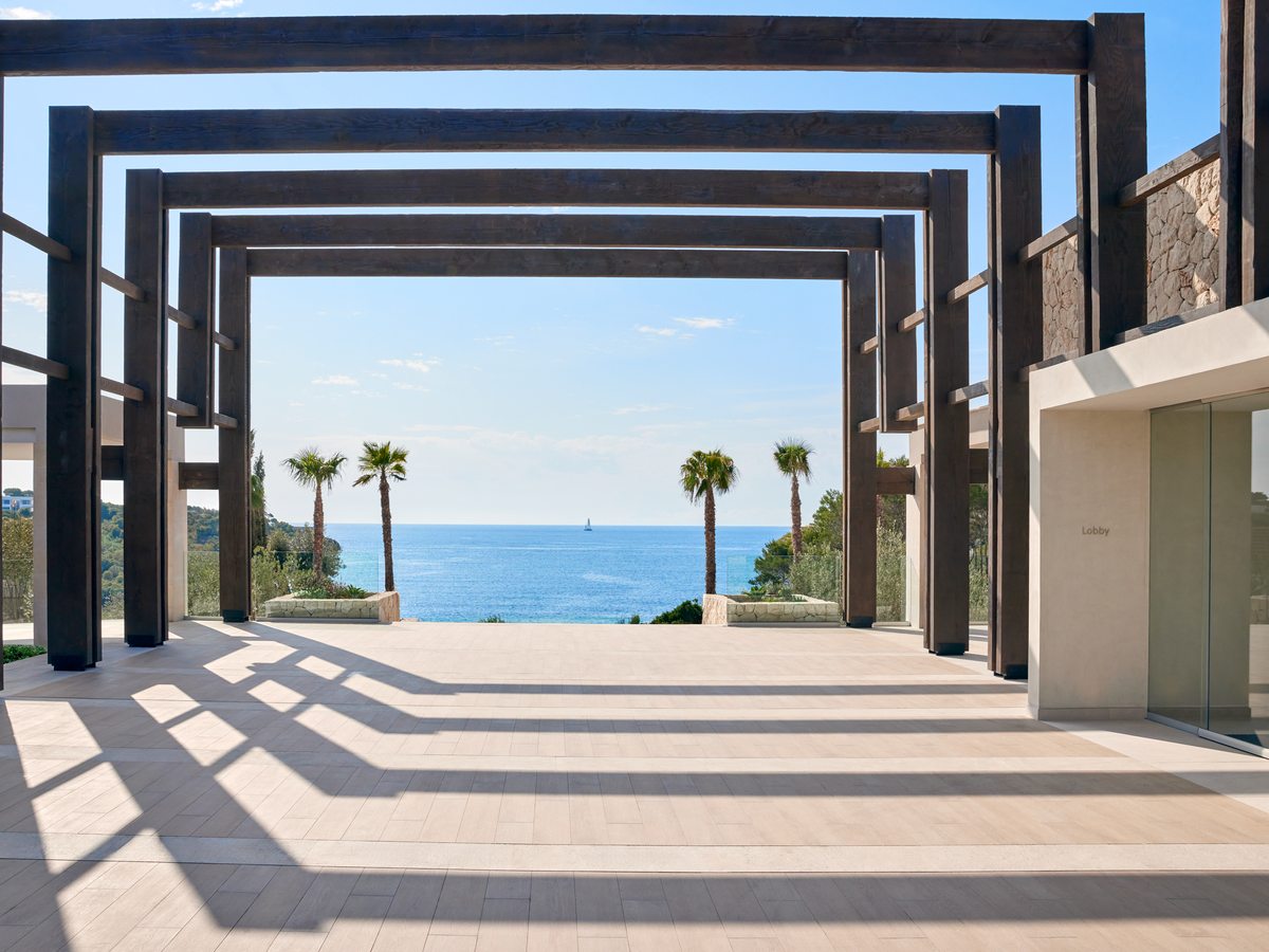 A run of tall timber-framed arches at the resort entrance over a paved walkway, with palms and the sea at the far end.