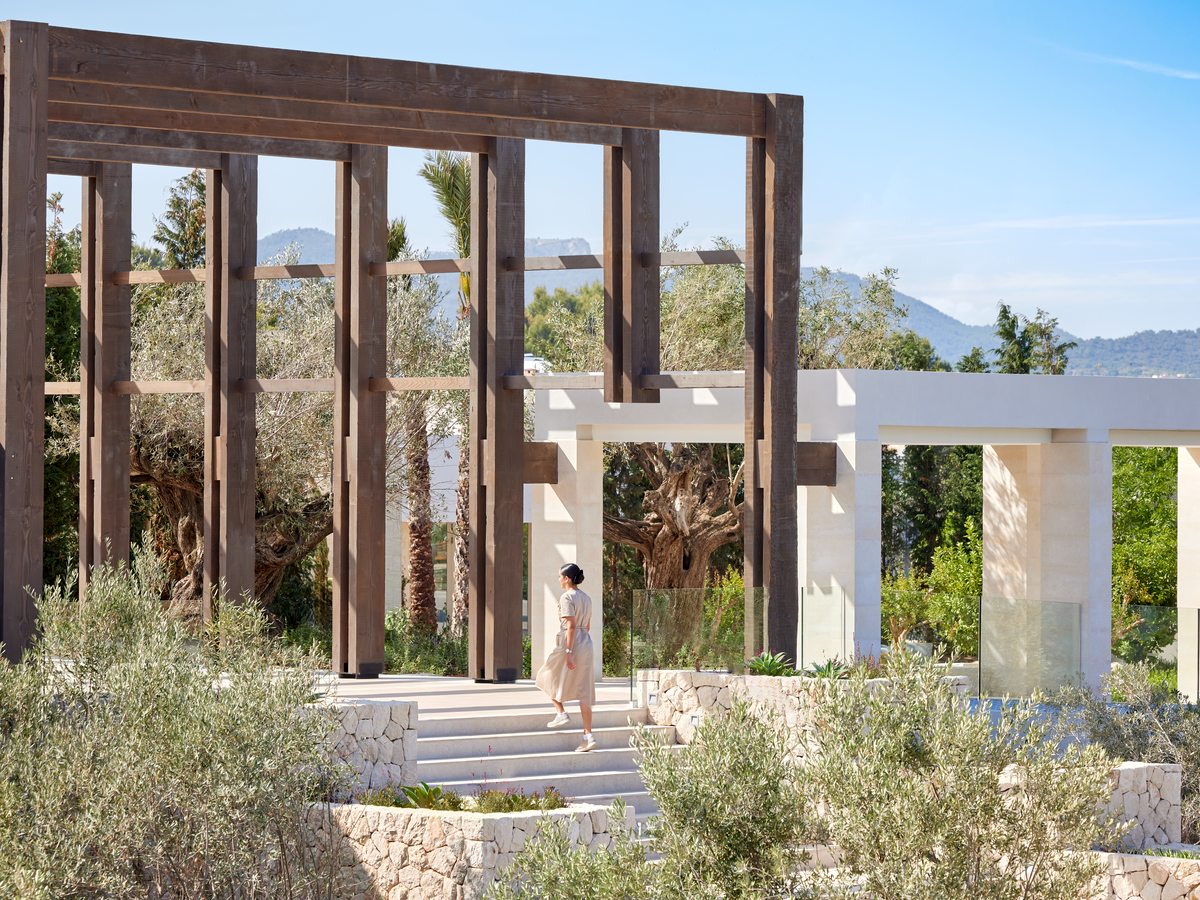 A colonnade of timber and stone frames over a stepped path through olive trees and low planting, with hills in the distance.