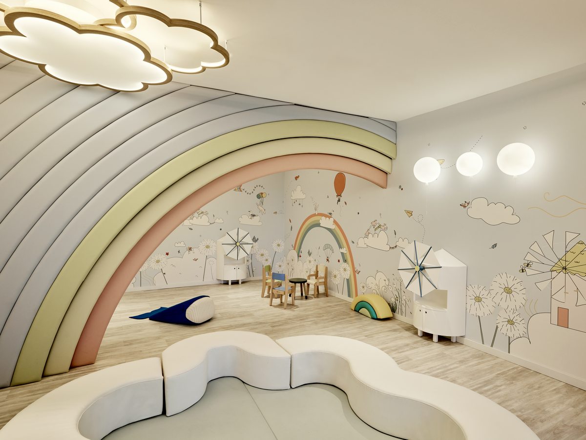 The baby centre playroom, with a painted rainbow arch over a low curved bench, cloud-shaped ceiling lights and small tables and chairs.