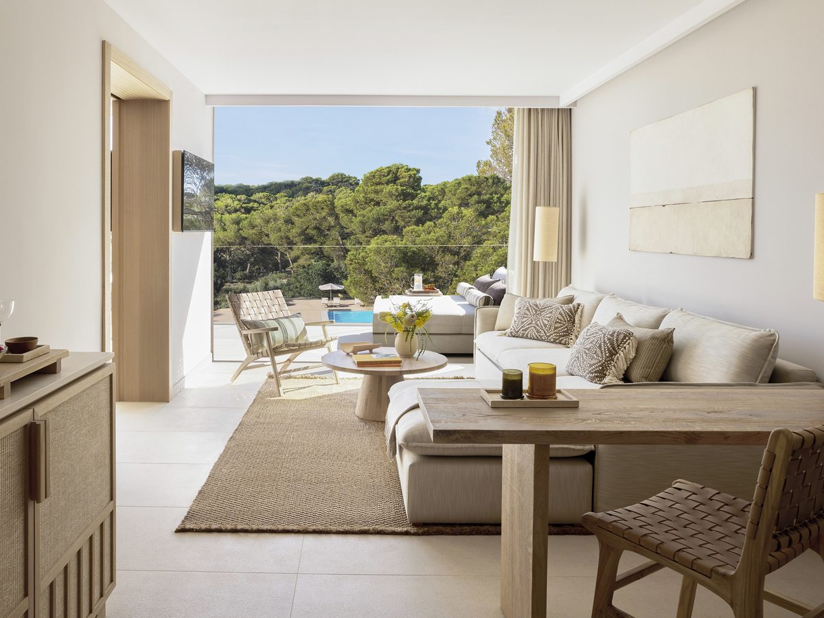 The living room of a One Bedroom Suite, with a sofa, a dining table and open doors onto a terrace, the pool and pines beyond.