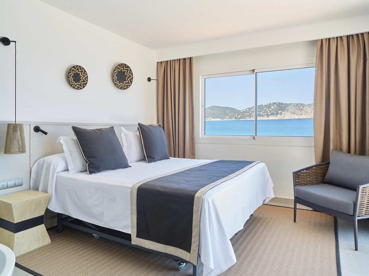 A double bed with a grey runner and a wicker chair beside a large window over the coast.