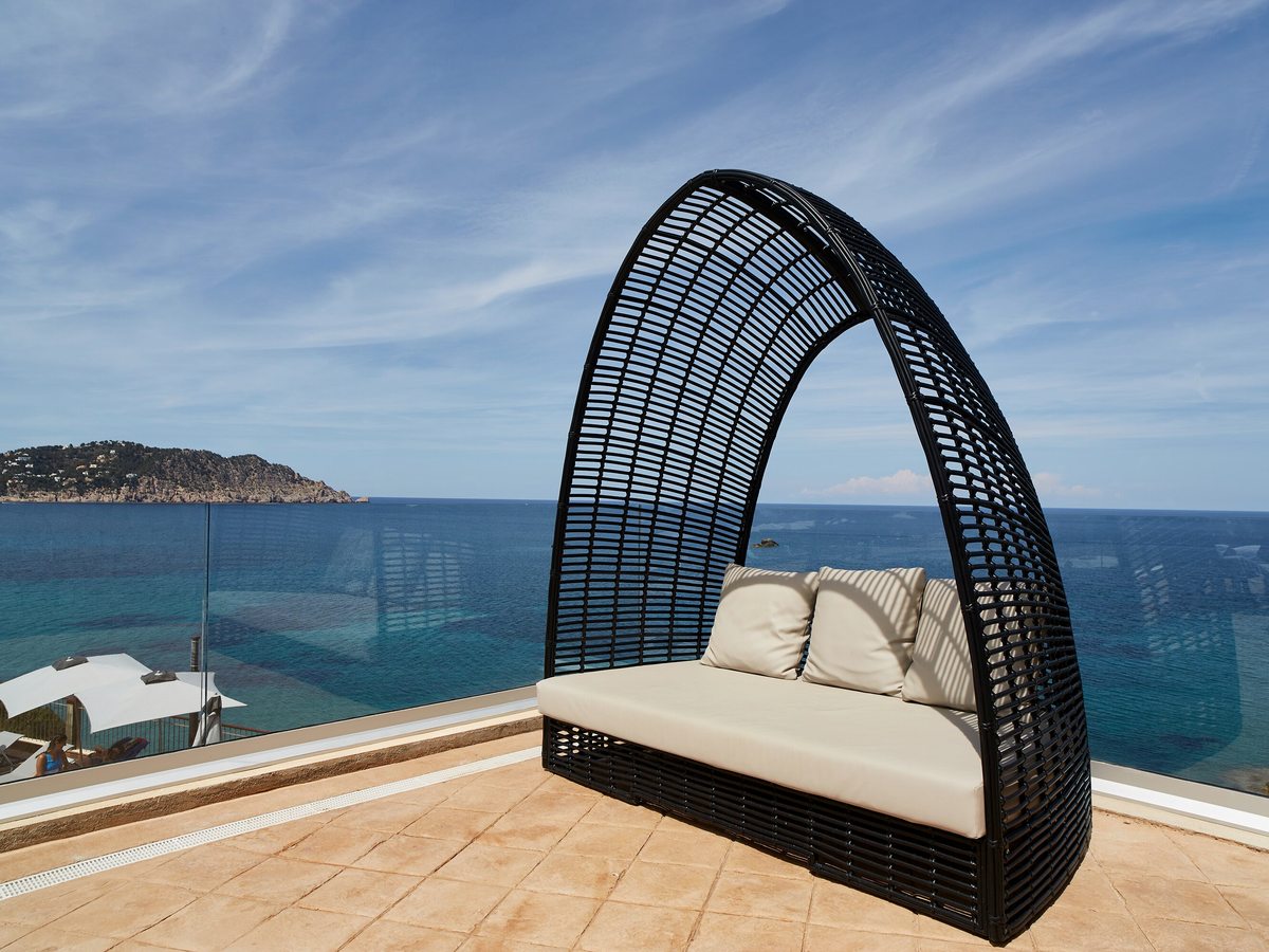 A woven daybed on a tiled terrace with a glass balustrade above the sea.