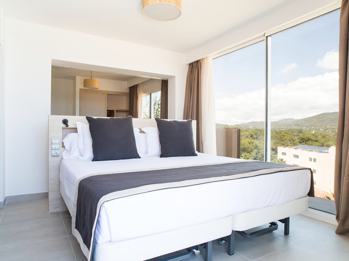 A double bed with dark cushions beside large windows facing the wooded hillside.