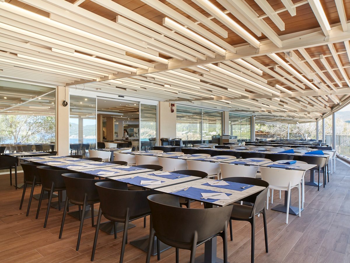 An indoor dining room with glass walls, blue placemats and a wood-slat ceiling beside the sea.