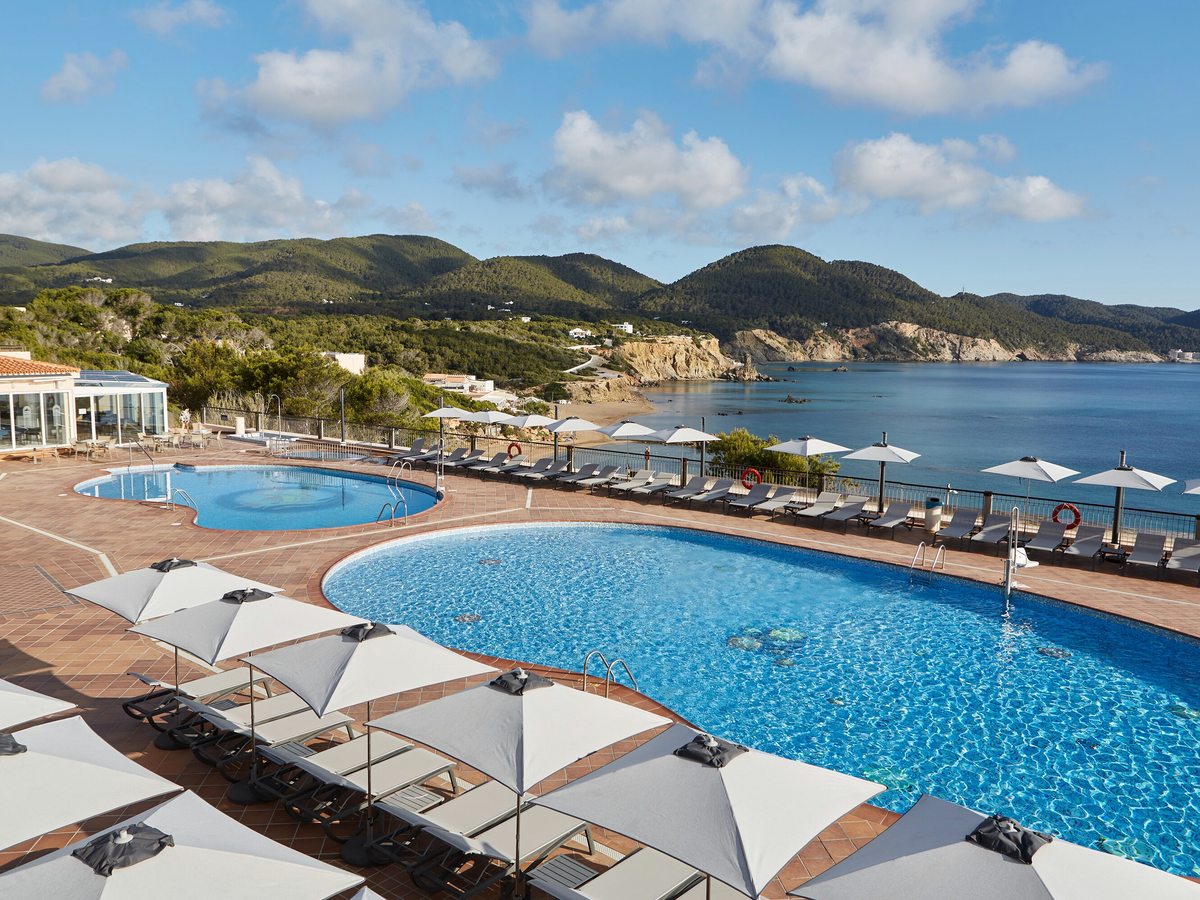 Two curved pools ringed by white parasols and loungers on a terrace above the bay and headland.