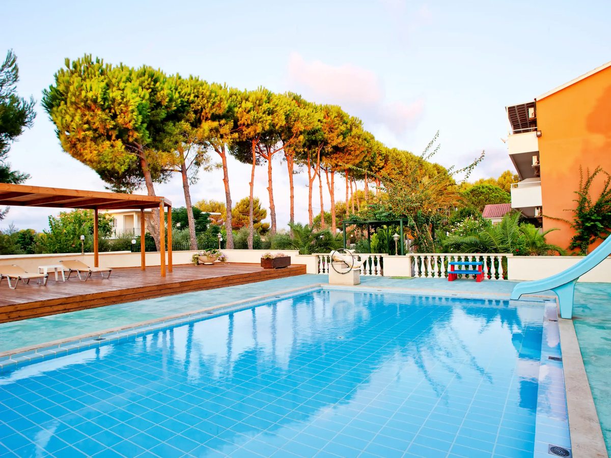A blue swimming pool edged with sun loungers and thatched umbrellas, backed by pine trees and an accommodation building.