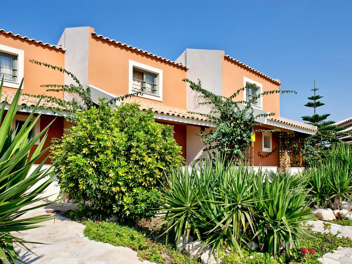 A peach accommodation building framed by tall greenery and red flowers under a clear blue sky.