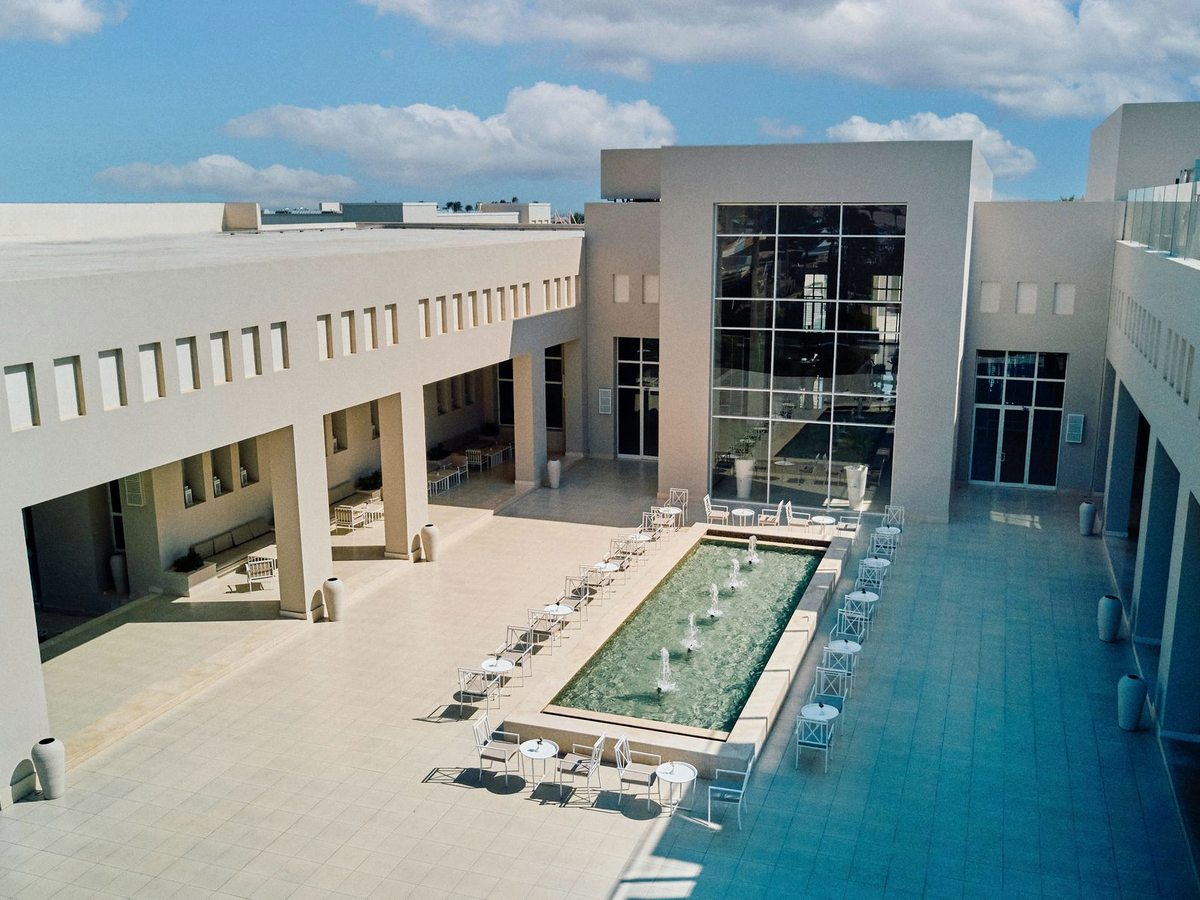 A long rectangular reflecting pool with fountain jets in the resort's cream-walled central courtyard, lined with white café tables and chairs below a tall glass-fronted block.