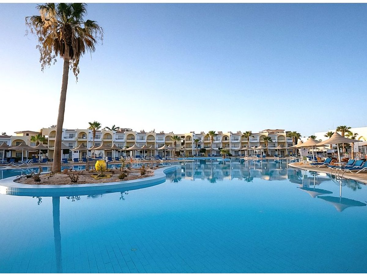 A large curved turquoise swimming pool with a small palm-planted island, straw parasols and blue sun loungers, backed by whitewashed two-storey accommodation blocks with arched balconies.