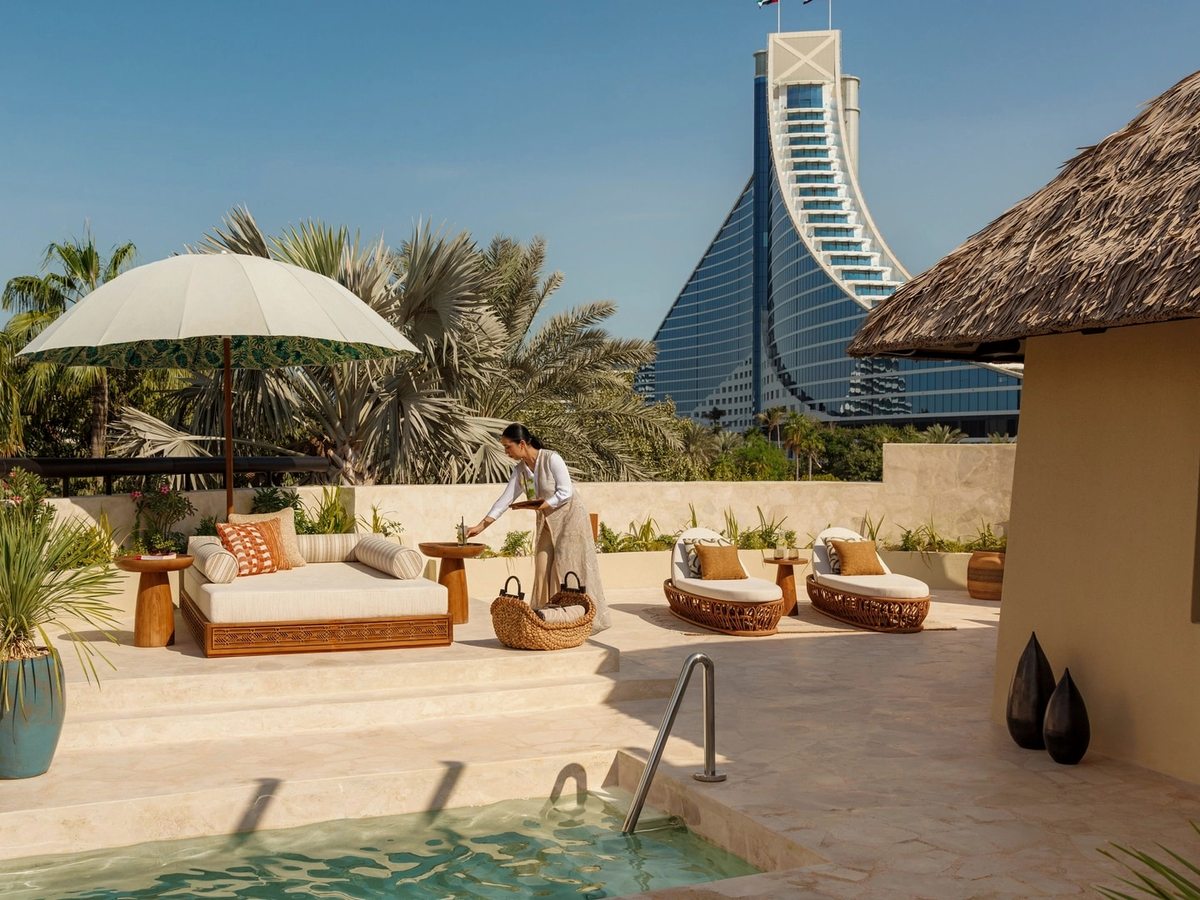 A villa terrace with a private plunge pool, a cushioned daybed under a parasol and wicker loungers, the wave-shaped hotel tower rising beyond the palms.