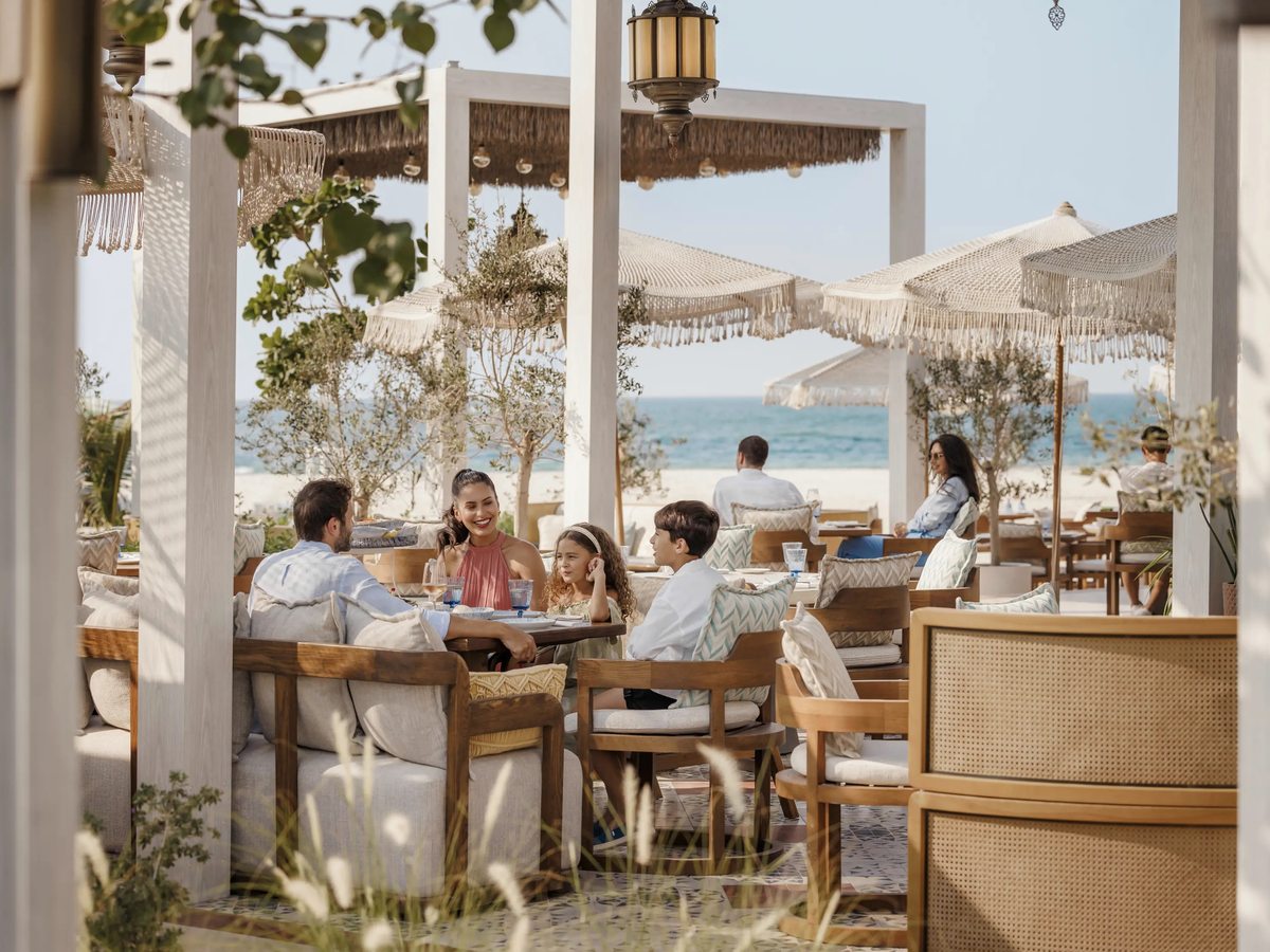 A beachfront restaurant terrace with white parasols and cushioned wicker chairs, a family at a table and the sea just beyond the sand.