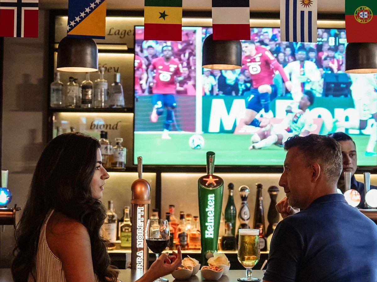 A bar with spirit shelves and beer taps, national flags strung overhead and a large screen showing football, two people talking at the counter.