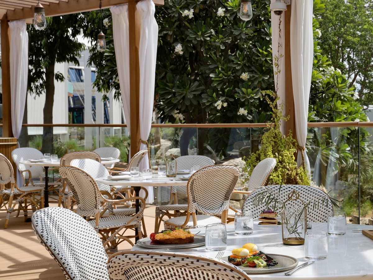 A shaded garden dining terrace under a timber pergola, with woven bistro chairs, white-clothed tables set with food and glasses, and dense greenery all around.
