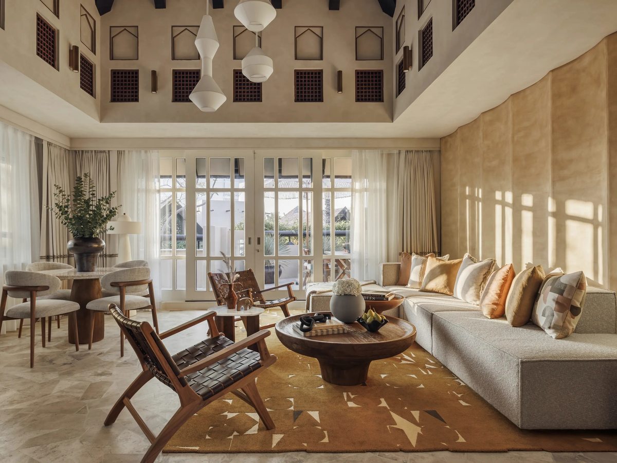 A villa living room with carved mashrabiya screens near the ceiling, a long sofa, woven leather chairs and French doors to a terrace.