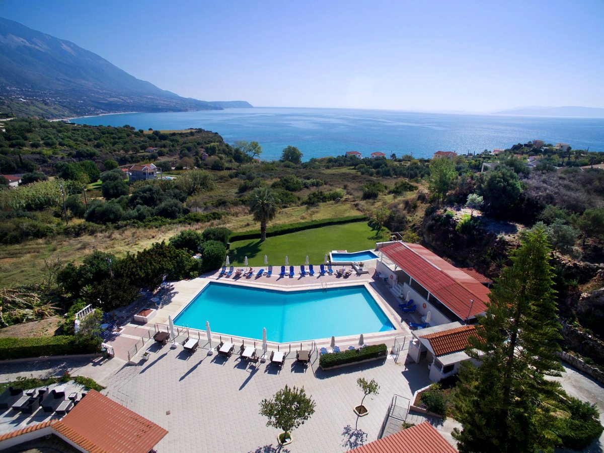 An aerial view of the hotel's large rectangular main pool and its paved sun terrace, with a smaller pool behind and the Ionian Sea and coastline beyond.