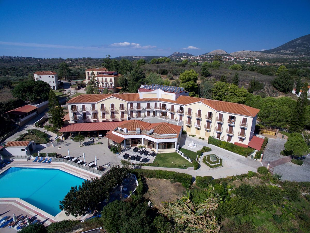 Aerial view of the hotel's cream buildings and a rectangular blue pool on a green hillside above the Ionian Sea.