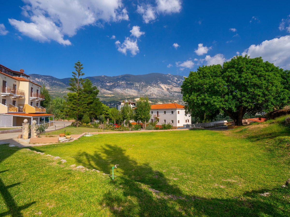 Green lawns and low hotel buildings in the foreground with the wooded slopes of Mount Ainos behind under a blue sky.