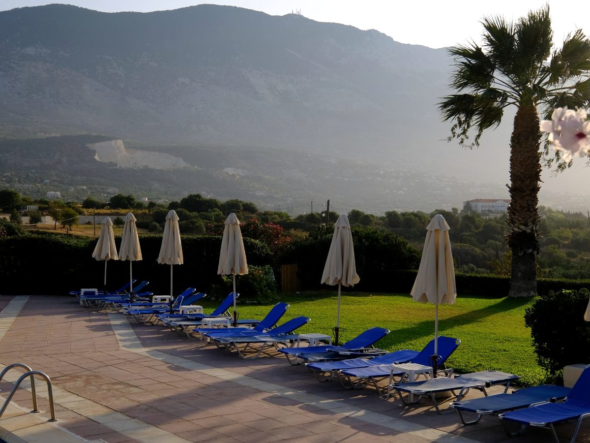 Rows of blue sun loungers with folded umbrellas on a paved terrace, with green lawn and mountains beyond.