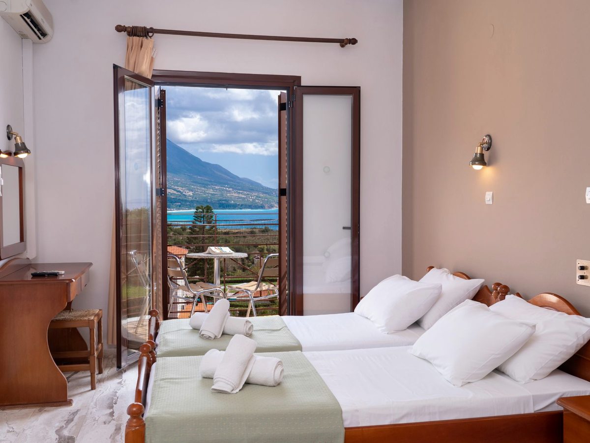 A room with two single beds, wooden furniture and open balcony doors showing a sea and mountain view.