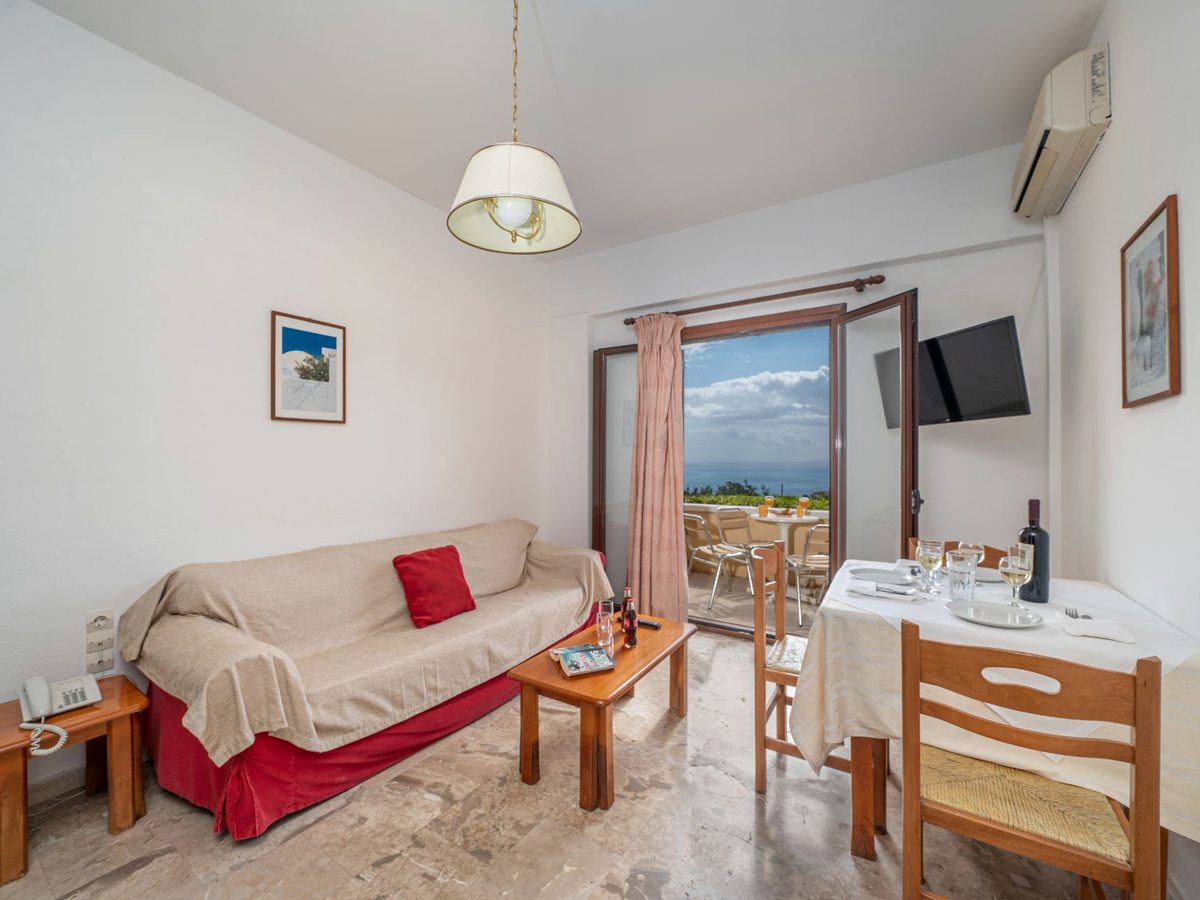 An apartment lounge with a sofa, coffee table, dining table and a wall-mounted TV, with a balcony beyond.