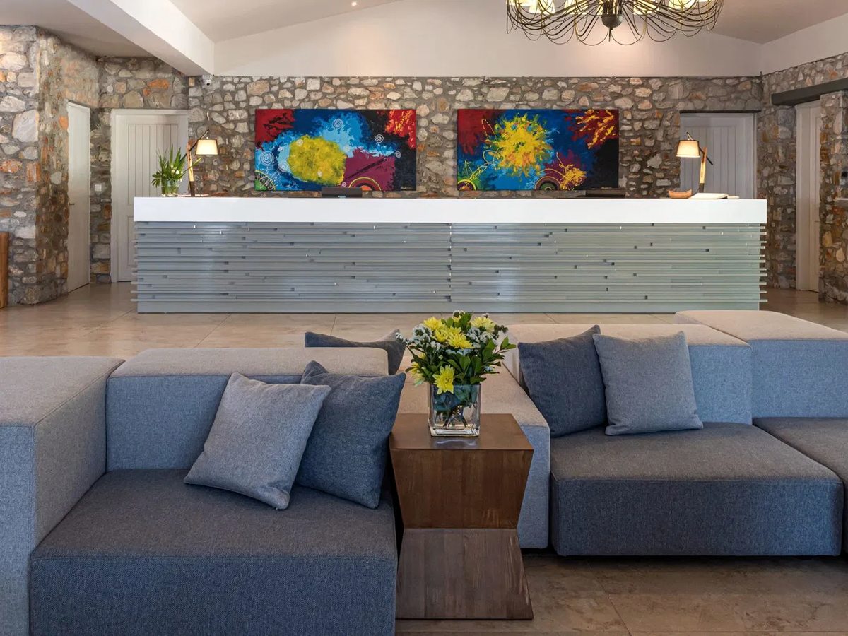 Hotel reception with a long white front desk, grey sofas, stone-clad walls, colourful abstract paintings and a chandelier over a tiled floor.