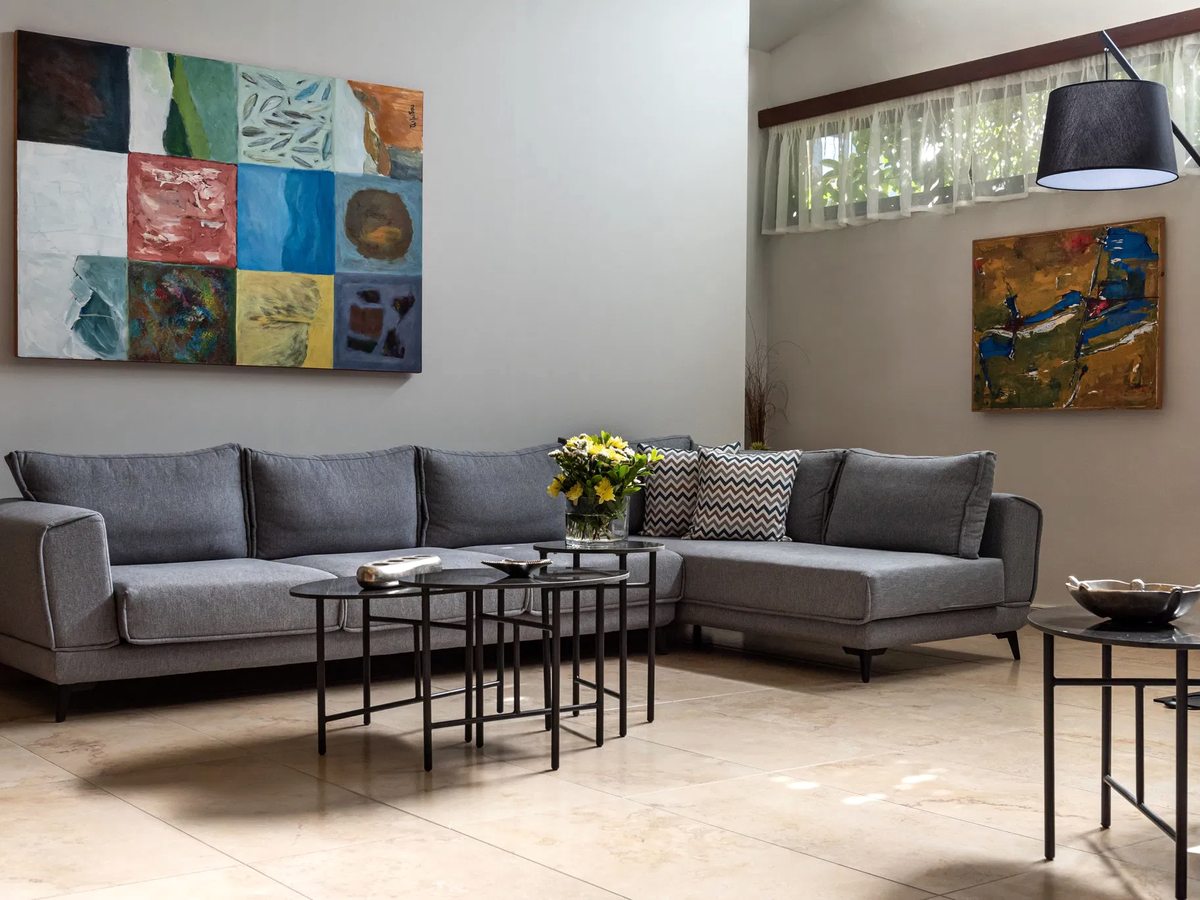 Lobby lounge with a grey sectional sofa, nested coffee tables, a floor lamp and colourful artwork below tall windows.