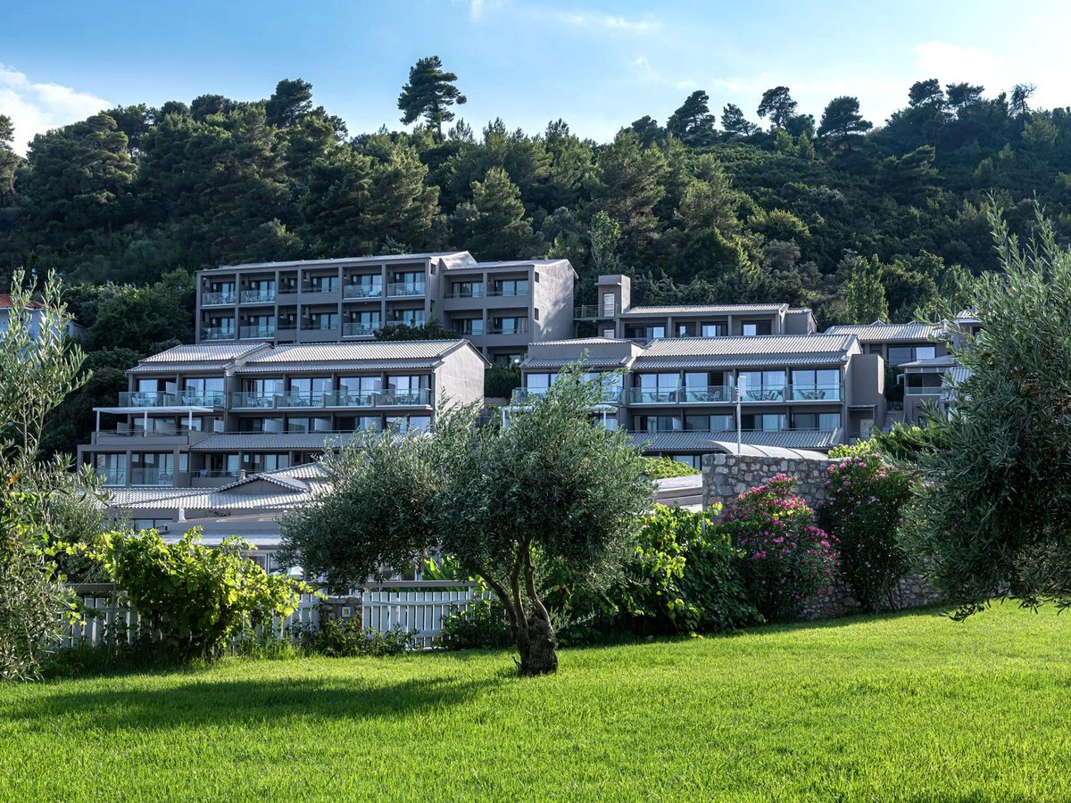 Resort gardens with a green lawn and olive trees in front of low-rise accommodation blocks with glass balconies on a wooded hillside.