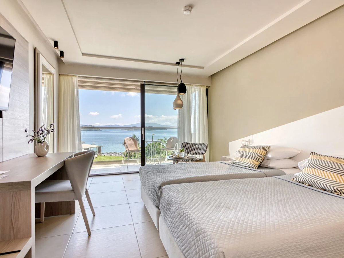 Guest room with twin beds pushed together, a desk and glass doors opening to a balcony with a sea view.
