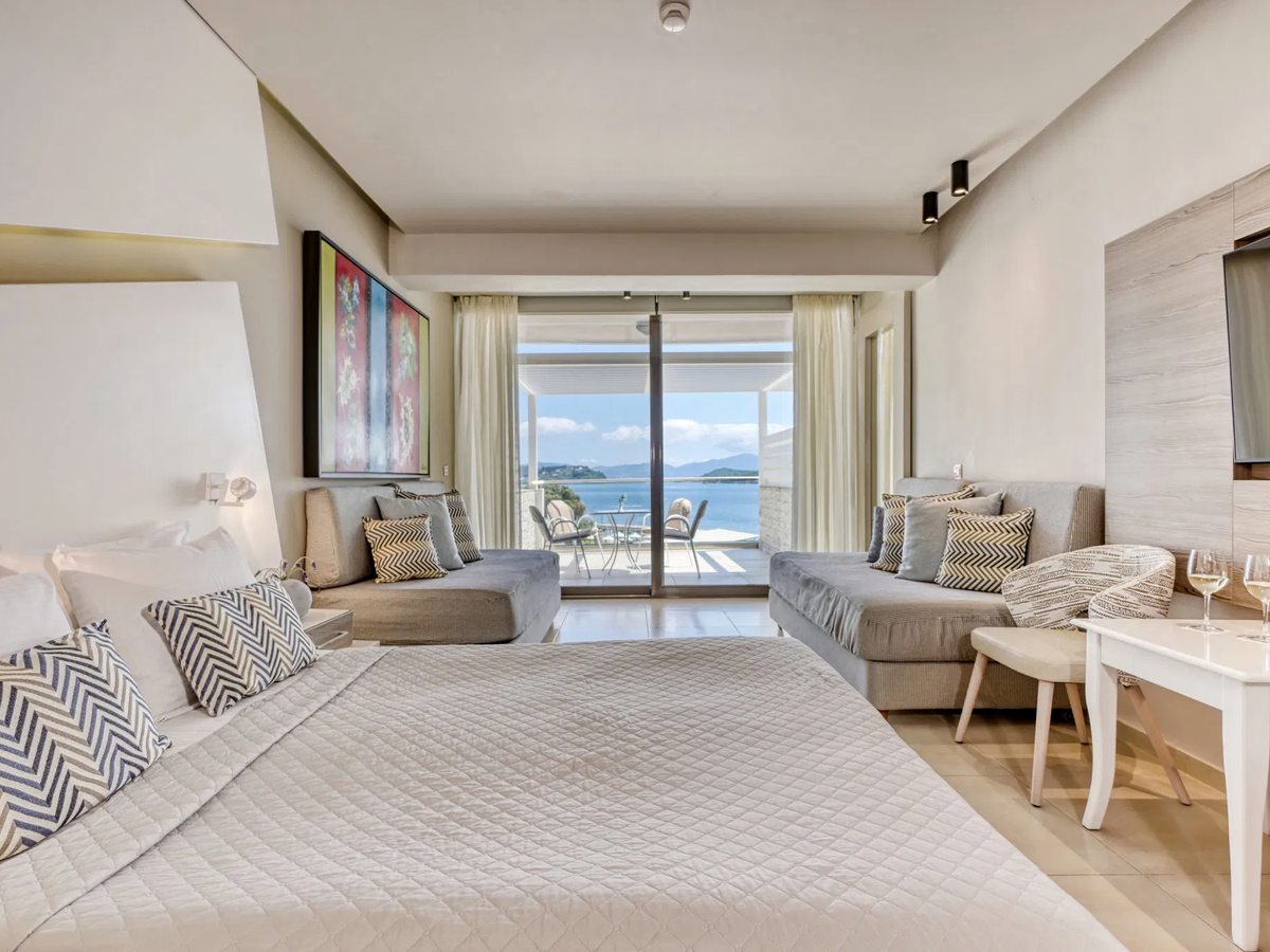 Spacious guest room with a double bed, a sofa and a desk, and glass doors to a sea-view balcony.