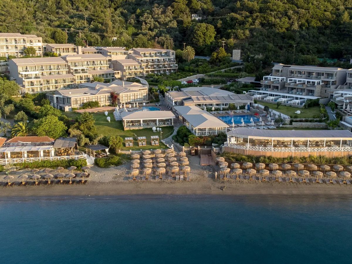 Aerial view of the resort's low-rise buildings, pool terraces and a sandy beach lined with straw parasols on a calm bay.